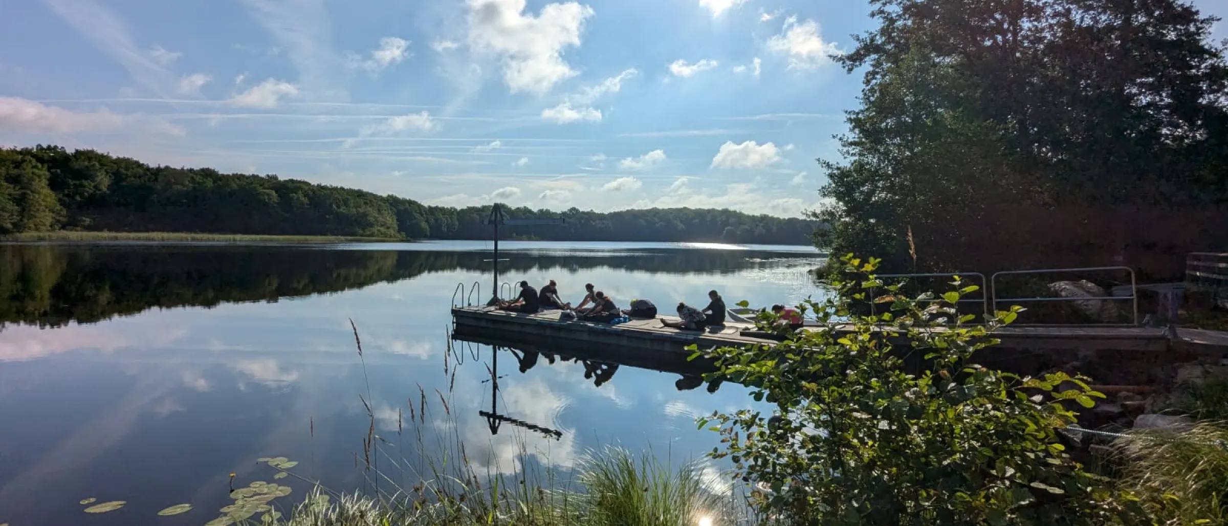 Das Bild zeigt einen See mit einem Steg, darauf sind Jugendliche zu sehen. Im Hintergrund sieht man einen blauen Himmel.